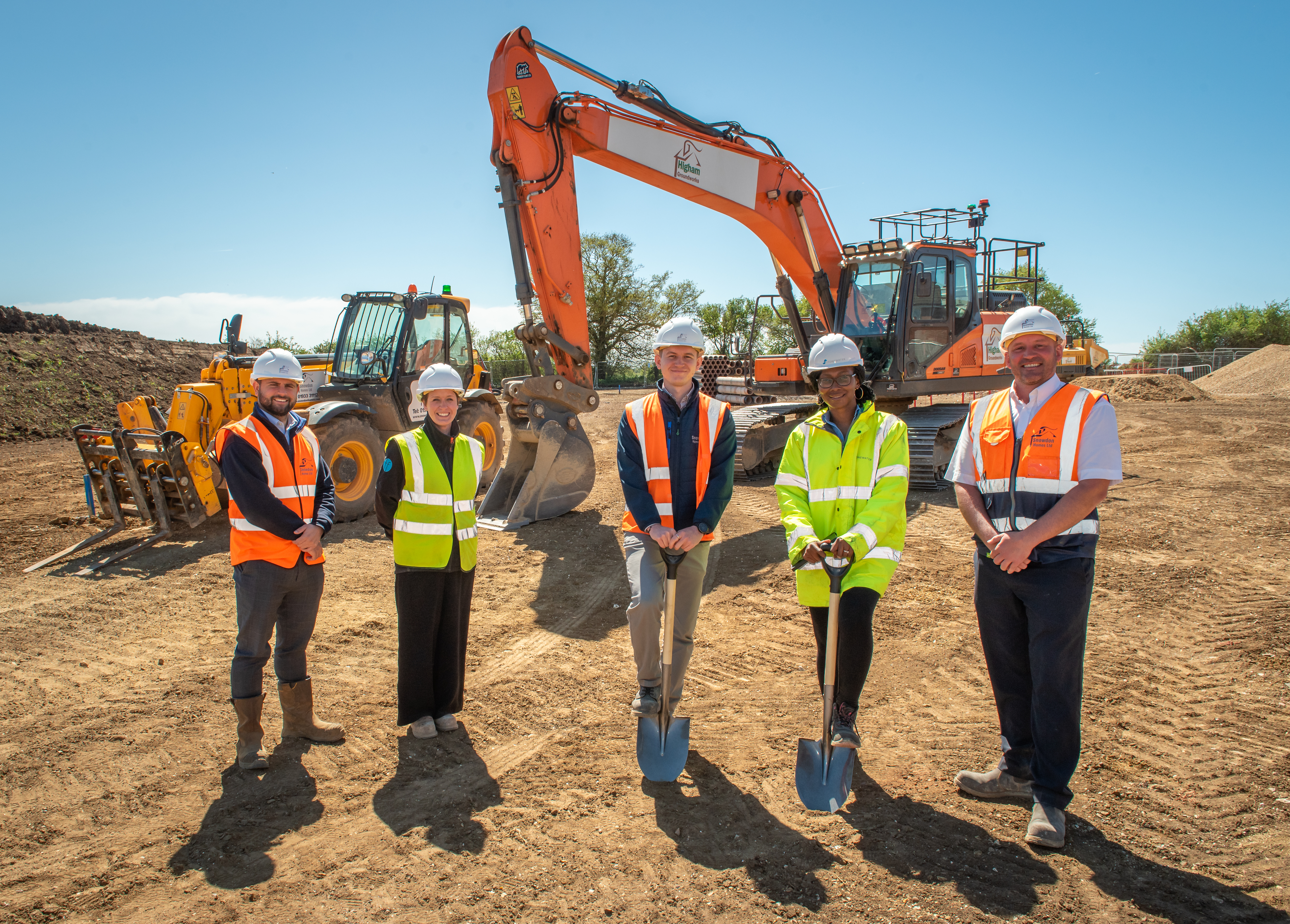 5 people including 2 with spades stand in front of a digger. They wear hard hats and hi vis vests.