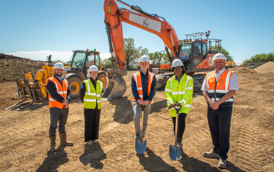 5 people including 2 with spades stand in front of a digger. They wear hard hats and hi vis vests.