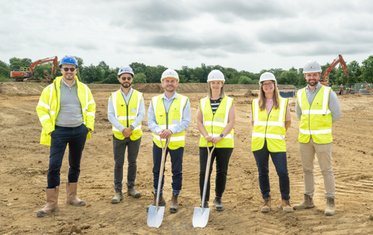 6 people wear PPE on a building site. Two hold spades as a ceremonial spade in the ground.