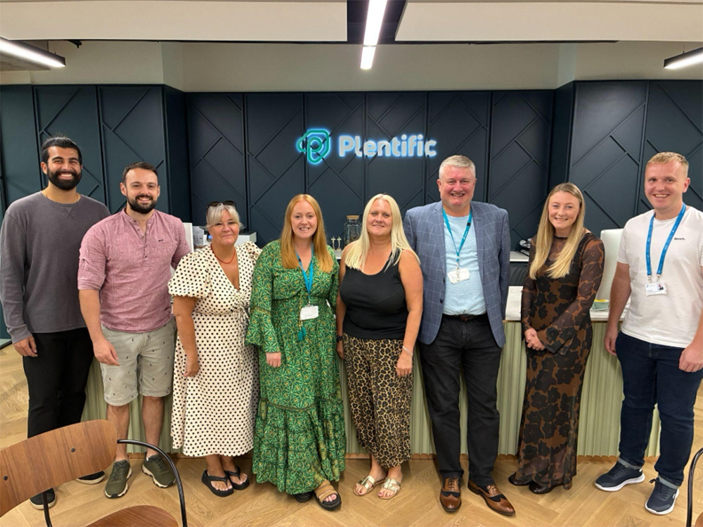 8 people stood smiling in front of a Plentific sign.