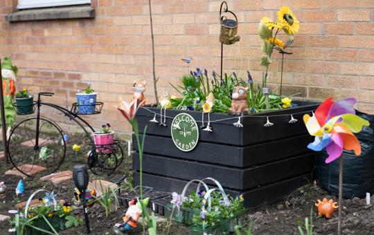 a black raised flower bed sits against a brick wall with seedlings and garden ornaments round it