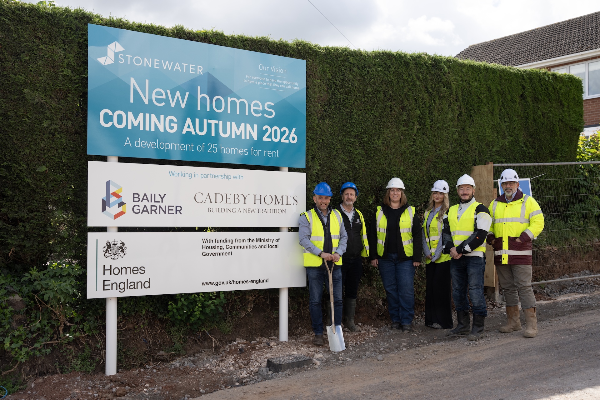 Row of people in hivis vests and hard hats beside a large sign about a new development
