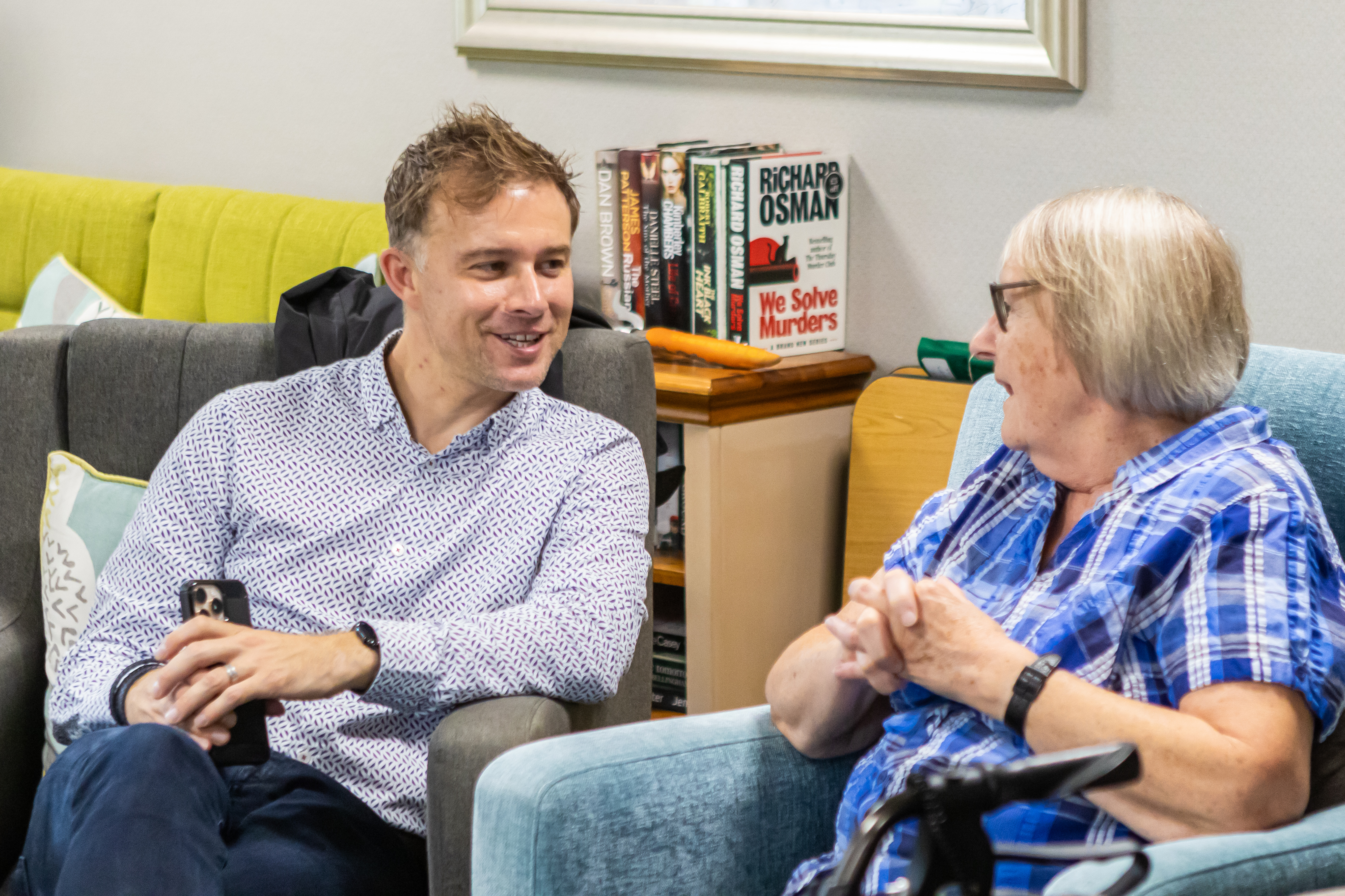 A man talking to a woman sat in a retirement living lounge with books on a bookcase behind
